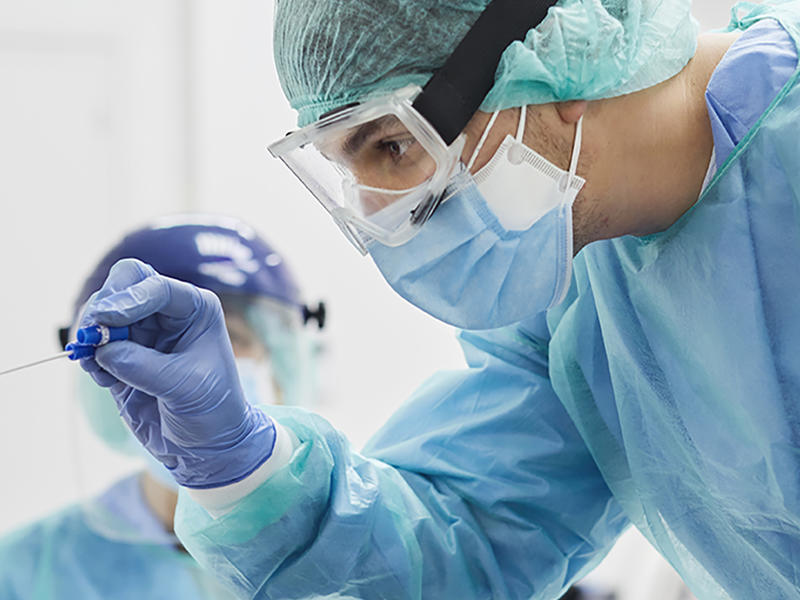 Clinician attired in full protective equipment including face shield examines throat of reclining patient.